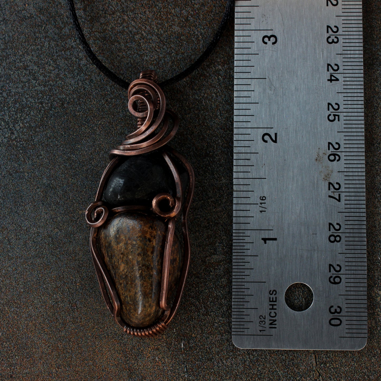 Copper pendant with stones on a dark surface next to a ruler for scale.
