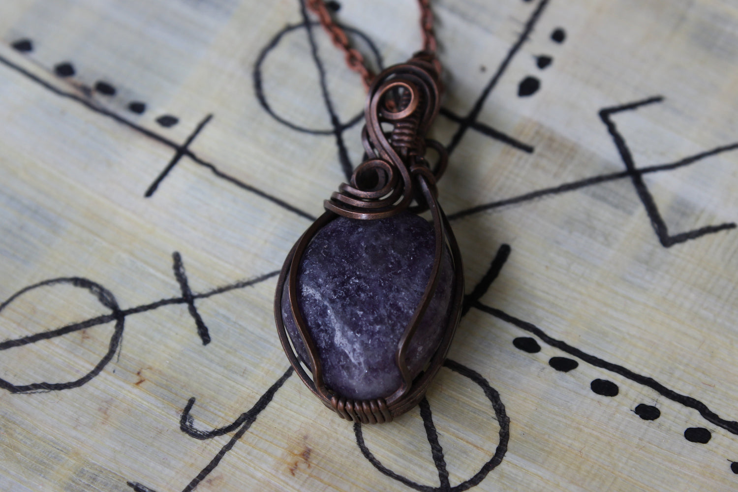 Purple Lepidolite stone pendant with oxidized copper wire wrapping on a papyrus surface with black line drawings.