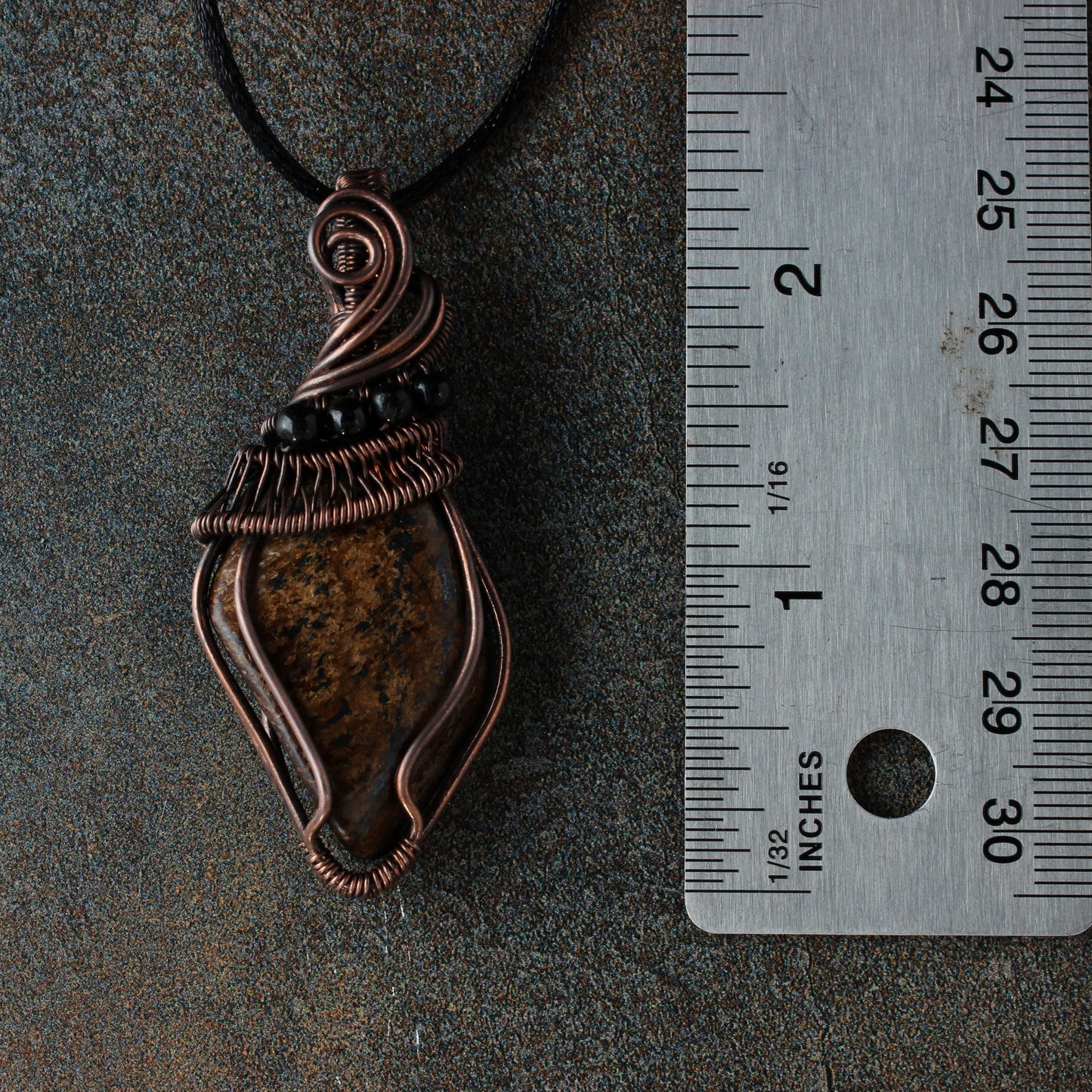 Copper pendant with black beads on a dark surface next to a ruler for scale.
