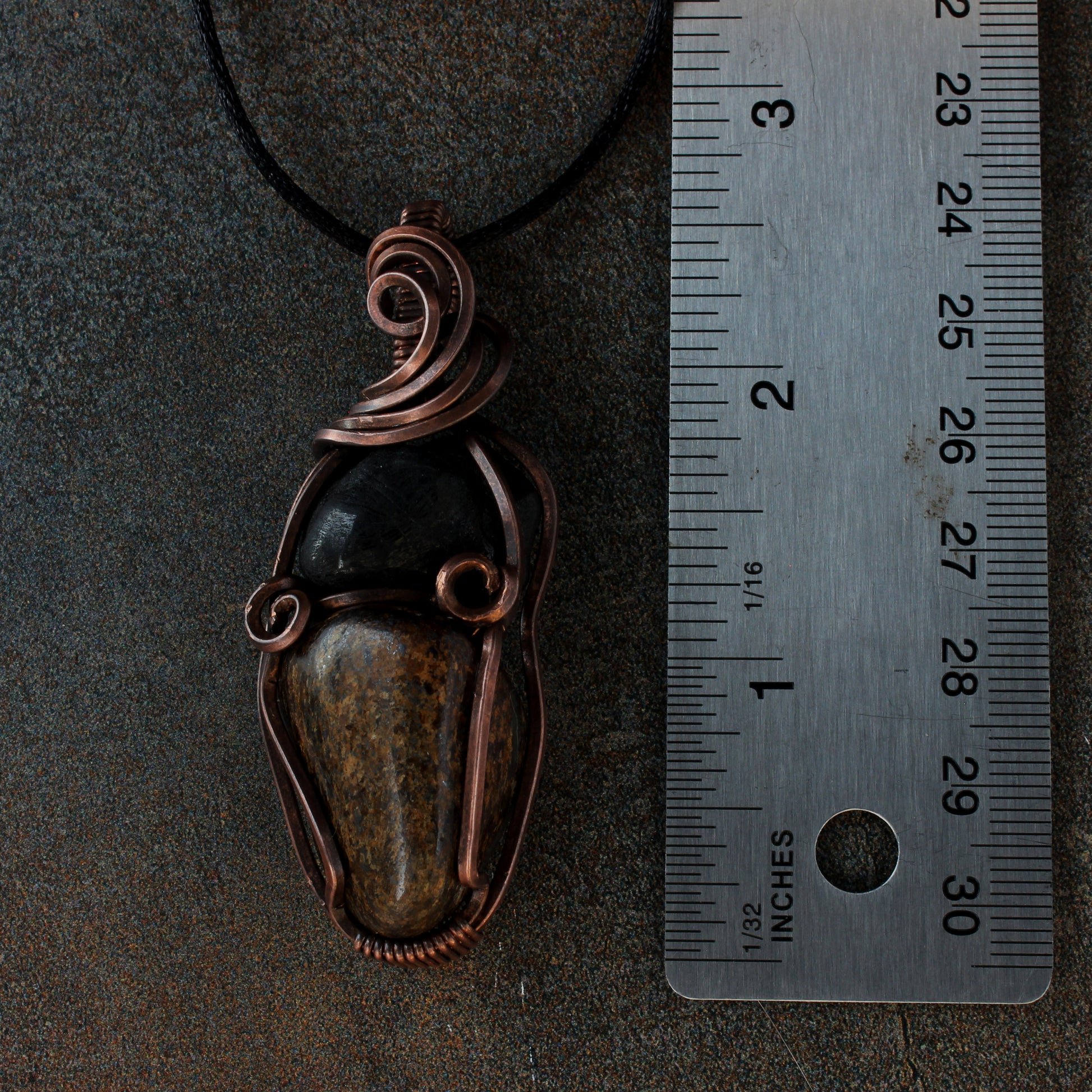 Copper pendant with stones on a dark surface next to a ruler for scale.