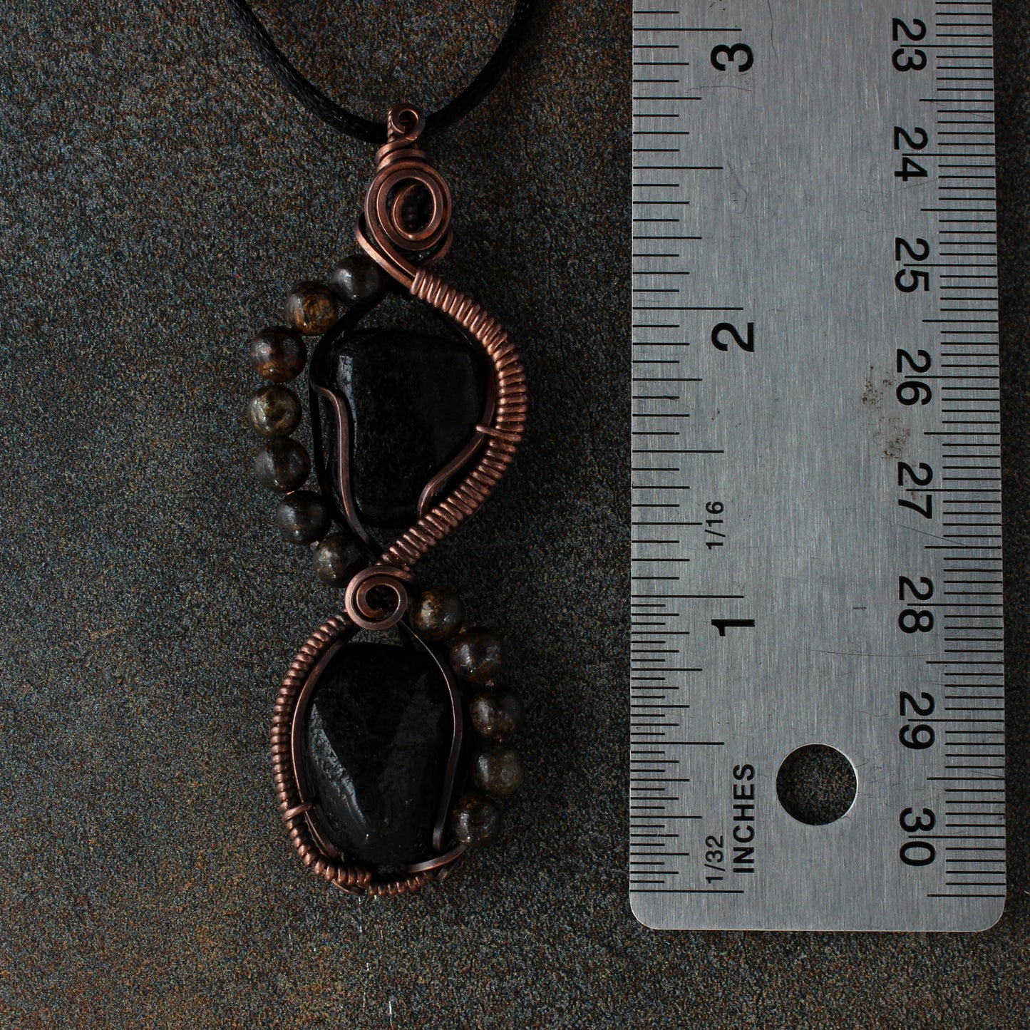 Copper and black stone pendant with a ruler for scale on a dark background